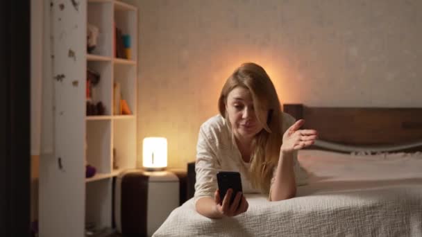 Jovem loira de pijama deitada na cama falando sobre smartphone telefone celular perplexo pelo estúpido interlocutor bobo - Filmagem, Vídeo