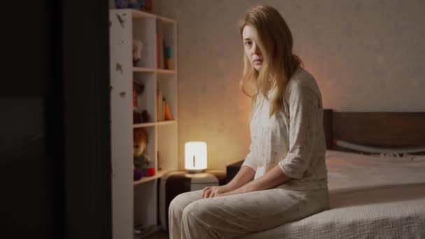 Zdenerwowany chory samotna dziewczyna w piżamie siedzi na kanapie w sypialni ze smutnym spojrzeniem w aparacie - Materiał filmowy, wideo