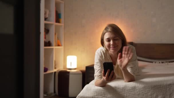 Boldog mosolygós szőke nő pizsamában feküdt az ágyon kényelmes otthon, hogy videohívás okostelefonon keresztül mobiltelefon kommunikálni mosolygós hétvégi öröm - Felvétel, videó