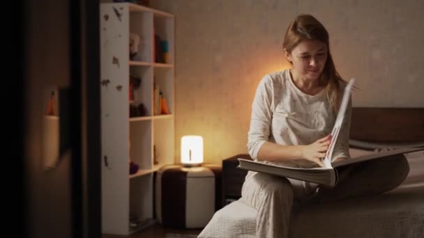 Boldog mosolygós szőke nő pizsamában ül az ágyon kényelmes kényelmes hálószobában nézi a fotóalbumot fényképekkel - Felvétel, videó