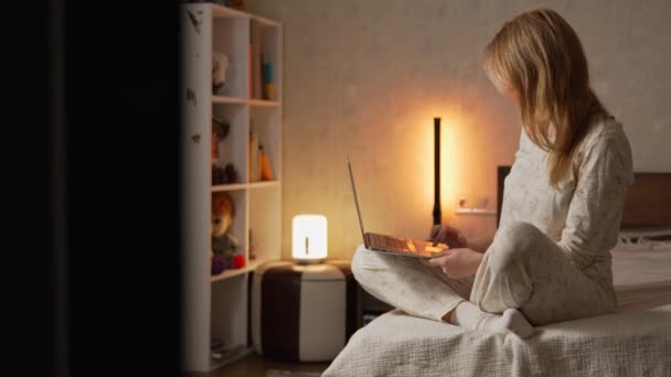 Γυναίκα με πιτζάμες κάθεται στο κρεβάτι σε ένα άνετο φωτεινό υπνοδωμάτιο και εργάζεται σε ένα φορητό υπολογιστή. Εργασία από το σπίτι έννοια - Πλάνα, βίντεο