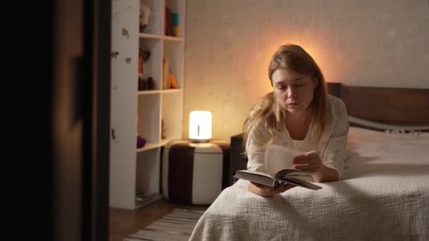 Mujer en pijama libro de lectura en el dormitorio pasa la página, acogedora iluminación tenue - Metraje, vídeo