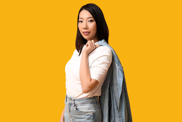 Belle jeune femme asiatique dans des vêtements en denim élégant sur fond jaune - Photo, image