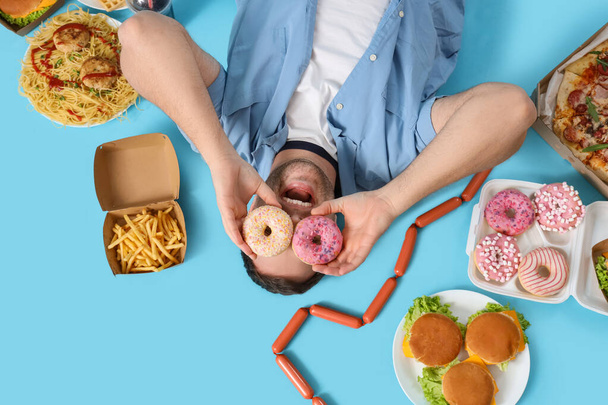 Молодой человек с нездоровой едой лежит на синем фоне, вид сверху. Концепция переедания - Фото, изображение