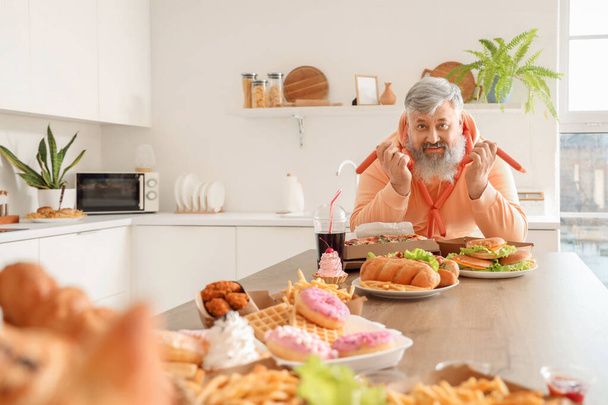 Uomo maturo sovrappeso a tavola pieno di cibo malsano in cucina. Concetto di eccesso alimentare - Foto, immagini