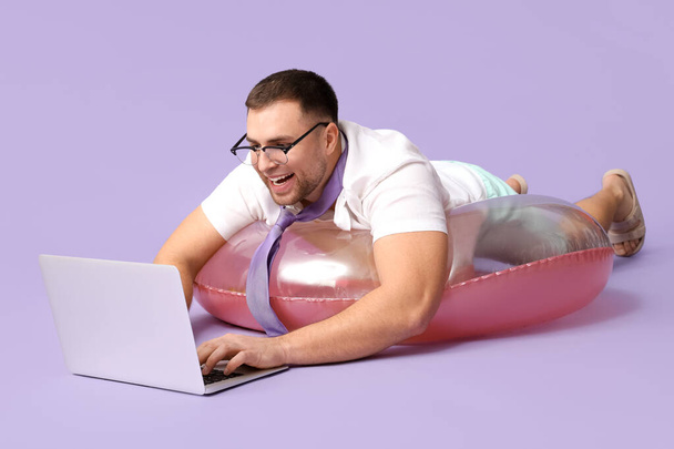 Szczęśliwy pracownik biurowy z laptopem leżącym na nadmuchiwanym pierścieniu na tle liliowym - Zdjęcie, obraz