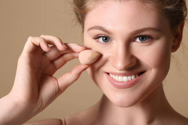 mooie jonge vrouw holding make-up spons met stichting op bruine achtergrond - Foto, afbeelding