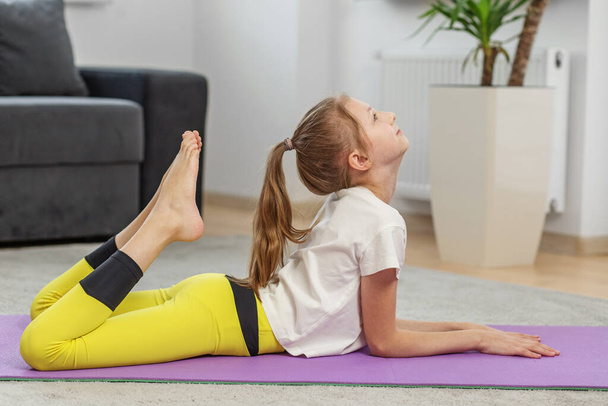 Mladá dívka provádí komplexní jóga stretch, zvedá nohu k hlavě na fialové podložce, ukazuje flexibilitu a dovednosti. - Fotografie, Obrázek