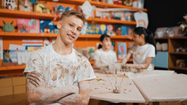 Smiling boy looking at camera and crossing arm with confident at workshop while diverse student having pottery class together. Happy caucasian student smile while pose with arm folded. Edification. - Photo, Image