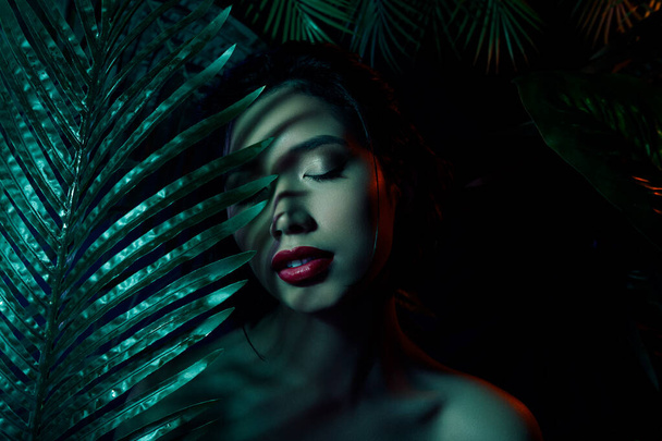 Foto de chica japonesa vietnamita sobre fondo oscuro cerrando los ojos se esconden detrás de palmeras exóticas. - Foto, imagen