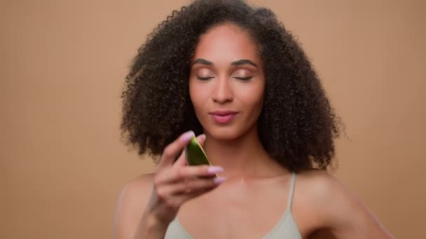 Afro-Amerikaans mooi gelukkig glimlachen mooi vrouw meisje vrouwelijk model met krullend haar in studio beige achtergrond houden half avocado vers groente gezond voeding eten bijten dieet gezondheidszorg voedsel - Video