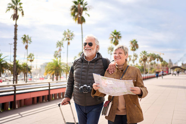 Couple of old Caucasian tourist walking hugging each other with their luggage facing forward. Smiling pensioner people enjoying their journeys and leisure time on a sunny winter day - Photo, Image