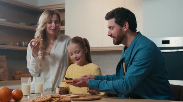 Ευτυχισμένος χαμογελαστός Καυκάσιος οικογένεια γονείς με παιδί κόρη τρώνε προετοιμάσει το πρωινό μαζί στην κουζίνα στο σπίτι απολαύσετε παράδοση βιολογικών τροφίμων τρώει σοκολάτα σάντουιτς μητέρα, ο πατέρας κοριτσάκι - Πλάνα, βίντεο