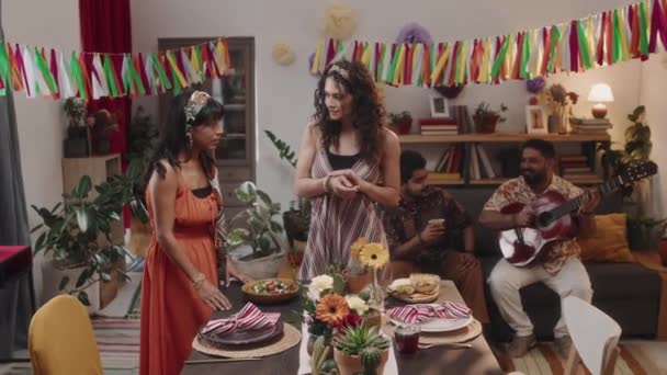 Teljes zoom felvétel két fiatal mexikói nőről sálakkal, fejpántokkal, fényes ruhákban, amint az asztalnál állnak ünnepi étkezésre és csevegésre, a férjek ülnek és gitároznak. - Felvétel, videó