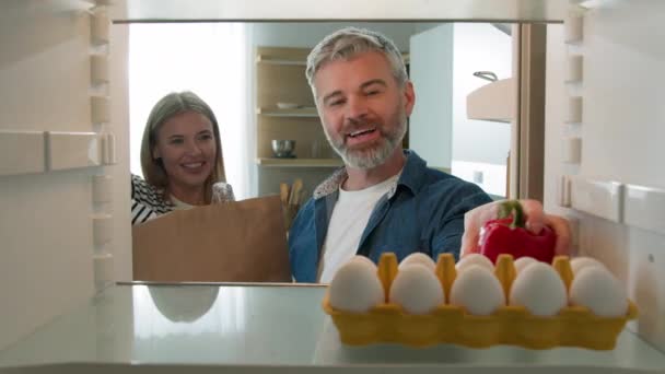 Взгляд изнутри холодильника Пара средних лет взрослые мужчина и женщина, муж и жена, беседующие распаковывая пакеты с продуктами питания, упаковывают овощи и полезные продукты в холодильник - Кадры, видео
