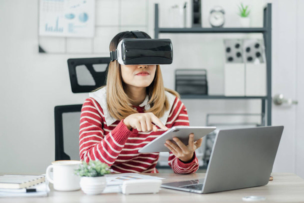 赤と白のストライプセーターの女性は,オフィスデスクで仮想現実のヘッドセットを着用しながらタブレットを使用しています.. - 写真・画像