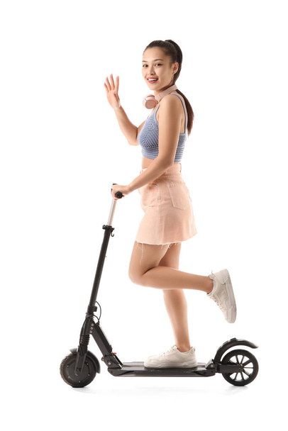 Felice giovane donna asiatica con le cuffie cavalcando moderno scooter elettrico calcio su sfondo bianco - Foto, immagini