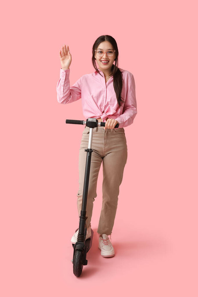 Όμορφη νεαρή Ασιάτισσα γυναίκα με μοντέρνο ηλεκτρικό πατίνι σε ροζ φόντο - Φωτογραφία, εικόνα