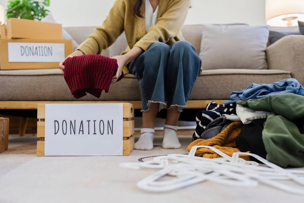 Jovens mulheres asiáticas se sentam na sala de estar classificando roupas para doação em uma caixa de doação roupas de segunda mão. Doar conceito. - Foto, Imagem