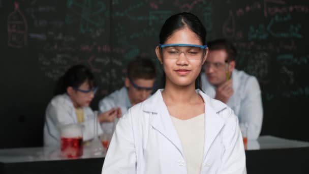 High-School-Mädchen blickt in die Kamera, während Menschen im Labor experimentieren. Junge süße Studentin posiert vor der Kamera, während sie an einer Tafel mit chemischer Theorie mit verschwommenem Hintergrund steht. Erbauung - Filmmaterial, Video