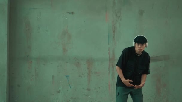 Jóképű hipster gyakorló break tánc közben zenét hallgat fejhallgató cement háttérrel. Jóképű ázsiai utcai táncos robottáncol. Kültéri sport 2024. Modern tánc. hiphop. - Felvétel, videó
