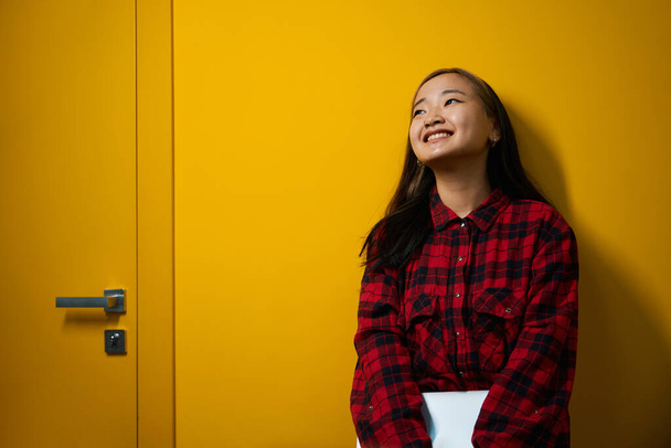 Nuori hymyilevä aasialainen IT-työntekijä, jolla on kannettava tietokone, seisoo seinän vieressä coworking-toimistossa. Käsitteen moderni freelance tai etätyötä - Valokuva, kuva