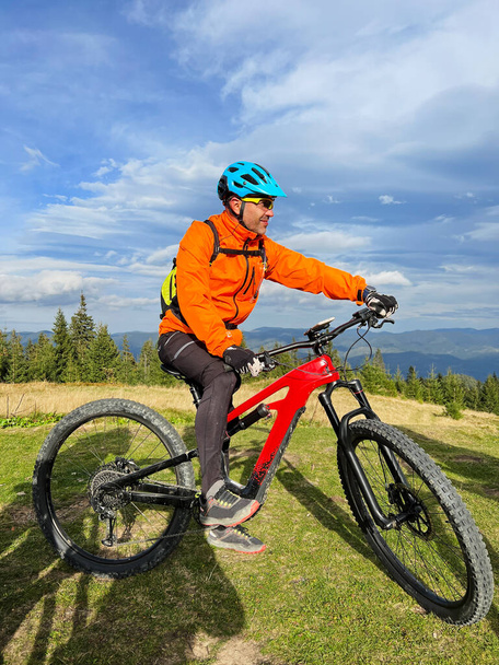 Kerékpárosok elektromos bicikliznek a szabadban, napsütéses napokon. Férfi turista pihen a füves dombon, élvezi a gyönyörű hegyi táj, visel sisak és hátizsák. Az aktív pihenés fogalma. - Fotó, kép