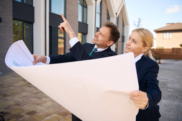 Обрезанное фото радостного архитектора-мужчины, показывающего дом женщине-клиенту, пока они держат план дома - Фото, изображение