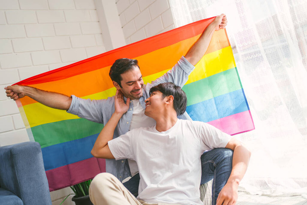 Happy caucasian and Asian LGBT couple, sitting on the sofa holding and waving rainbow LGBT Pride flag together in the living room at home. Diversity of LGBT relationships. A gay couple concept. - Fotó, kép