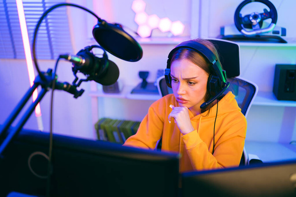 Młoda biała kobieta profesjonalny gracz w żółtej bluzie i słuchawki siedzieć na krześle z ciekawym nastroju, komputer, klawiatura, monitor, mikrofon. Przygotuj się na zawody, obsadę gameplay lub nagranie podcastu - Zdjęcie, obraz