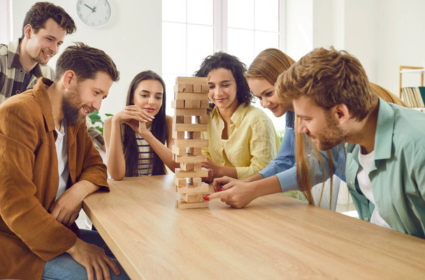 Group of a happy young friends guys and girls playing together with wooden building blocks at home sitting at the table enjoying time together. Home leisure and board games concept. - Fotó, kép