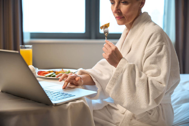 Zaostřená dospělá běloška snídá a ráno používá notebook na posteli u stolu v hotelovém pokoji. Koncept odpočinku, dovolené a cestování - Fotografie, Obrázek