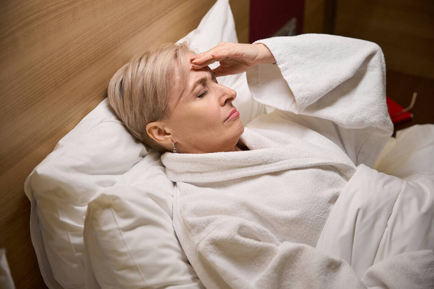 Εξαντλημένη ενήλικη καυκάσια γυναίκα φορώντας μπουρνούζι ξαπλωμένη και αγγίζοντας το κεφάλι της στο κρεβάτι στο δωμάτιο του ξενοδοχείου με φωτισμό. Έννοια της ανάπαυσης, των διακοπών και των ταξιδιών - Φωτογραφία, εικόνα