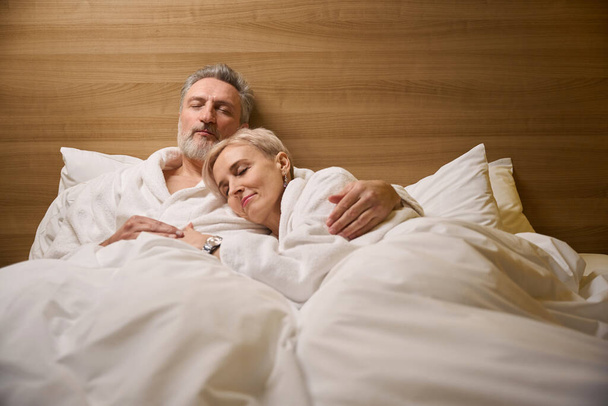 Coppia caucasica adulta che indossa accappatoi che abbracciano e dormono sul letto in camera d'albergo con illuminazione. Concetto di riposo, vacanza e viaggio. Idea di relazione - Foto, immagini