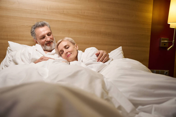Volwassen blanke man knuffelt zijn slapende vrouw op bed in de hotelkamer met verlichting. Concept van rust, vakantie en reizen. Relatiegedachte - Foto, afbeelding
