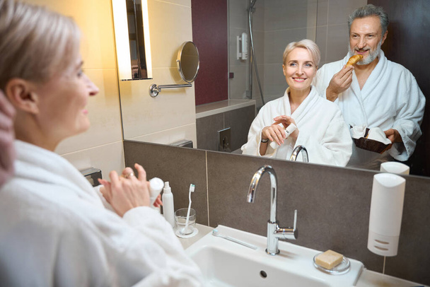 Smiling adult caucasian woman with cosmetic cream and man with croissants looking at themselves in mirror in bathroom at morning time. Concept of morning procedures and hygiene - Photo, Image