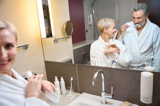Lustro odbicie uśmiechniętej białej pary dorosłych kobiet z kremem kosmetycznym patrząc na mężczyznę myjącego zęby szczoteczką do zębów w łazience w godzinach porannych. Pojęcie procedur porannych i higieny - Zdjęcie, obraz
