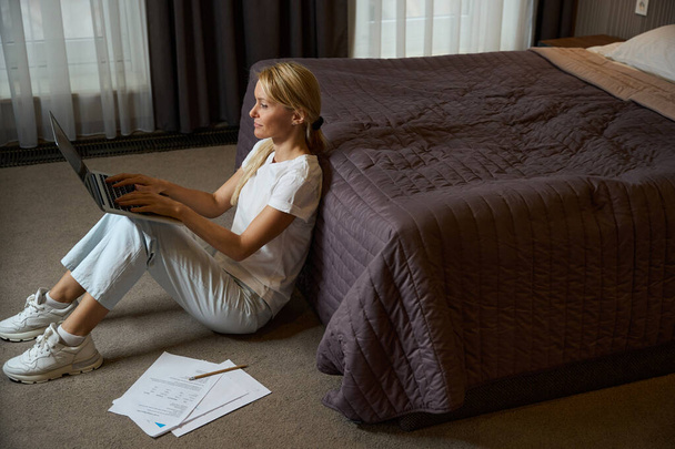 Koncentrált nő gépel hordozható számítógépen, miközben ül a szőnyegen az ágy mellett a szállodai szobában - Fotó, kép