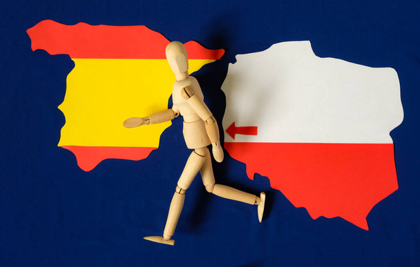 Ξύλινη ανθρώπινη κούκλα τρέχει. Πολωνικός χάρτης Ισπανικός χάρτης. Βέλος. Μετανάστευση πληθυσμού από την Πολωνία στην Ισπανία. Μπλε φόντο της σημαίας της Ευρωπαϊκής Ένωσης. - Φωτογραφία, εικόνα
