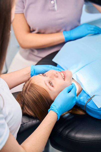 Paciente jovem sorridente colocada em uma cadeira para procedimentos odontológicos, equipe médica trabalhando em luvas de proteção - Foto, Imagem