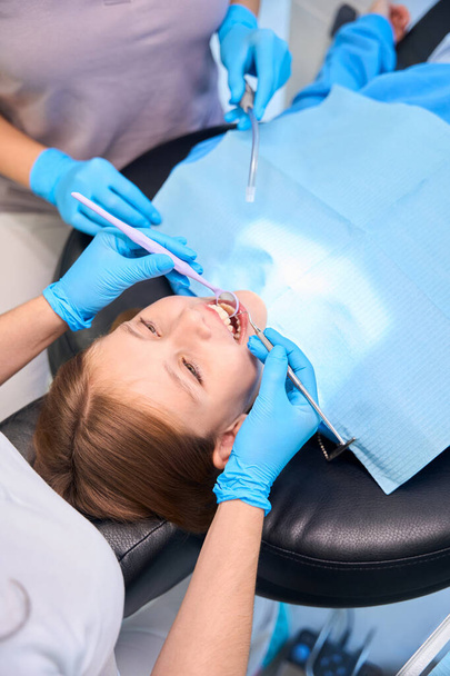 Diş hekimi koltuğunda oturan bir kız. Tıp personeli iş yerinde özel aletler kullanıyor. - Fotoğraf, Görsel