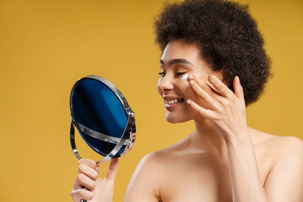 Красивая улыбающаяся афроамериканская молодая женщина держит и смотрит в зеркало, нанося увлажняющий крем на лицо, стоящее изолированным на желтом фоне. Концепция ухода за кожей, утренняя, косметическая процедура - Фото, изображение