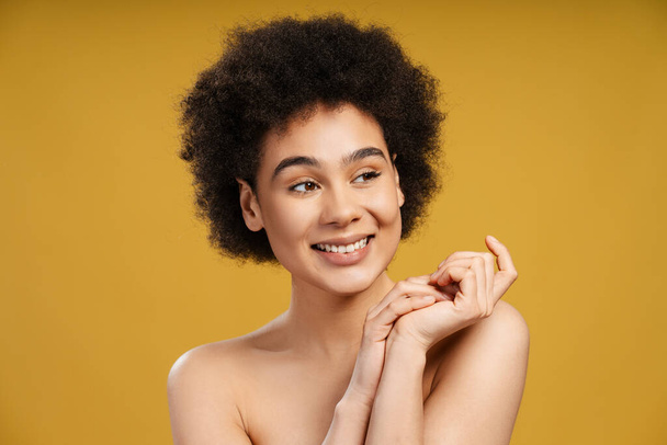 Mujer afroamericana sensual, confiada y sonriente con un peinado elegante mirando hacia otro lado. Hermosa modelo de moda posando aislada sobre fondo amarillo. Concepto de publicidad, belleza natural - Foto, Imagen
