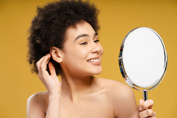 Attractive, happy African American woman holding and looking in mirror morning routine. Smiling young female after shower isolated on yellow background. Concept of hygiene, cosmetics procedure - Foto, afbeelding