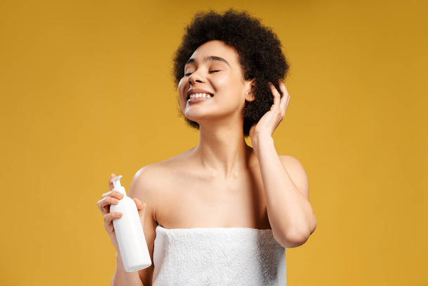 Hermosa modelo de moda, joven mujer afroamericana en toalla blanca sosteniendo botella con producto cosmético, maqueta con los ojos cerrados de pie aislado sobre fondo amarillo. Concepto de publicidad - Foto, imagen