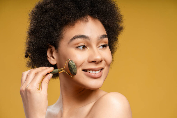 Attractive, happy African American woman with curly hair doing face massage with massage roller, looking away, standing isolated on yellow background. Concept of skin care, spa, anti aging - Photo, Image