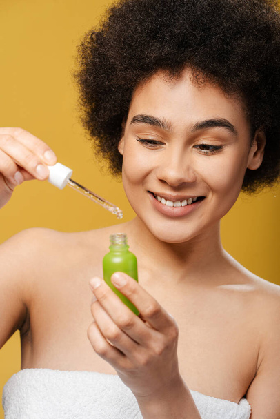 Zbliżenie atrakcyjna młoda Latynoska kobieta w białym ręczniku po prysznicu trzymając zieloną butelkę z serum, stojąc odizolowany na żółtym tle. Koncepcja pielęgnacji skóry, kosmetyka - Zdjęcie, obraz