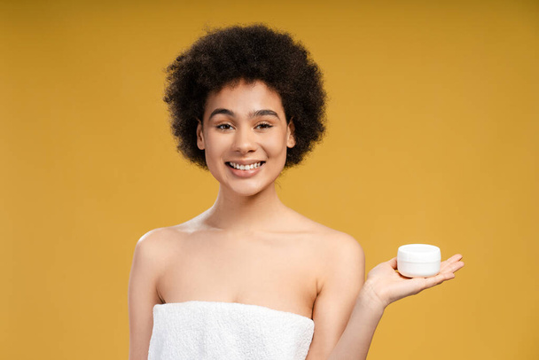 Gyönyörű afro-amerikai nő göndör hajjal, fehér üveget tart tejszínnel, fehér törölközőt visel, és a sárga háttéren álló kamerát nézi. Bőrápolás, kozmetikai termék koncepció - Fotó, kép