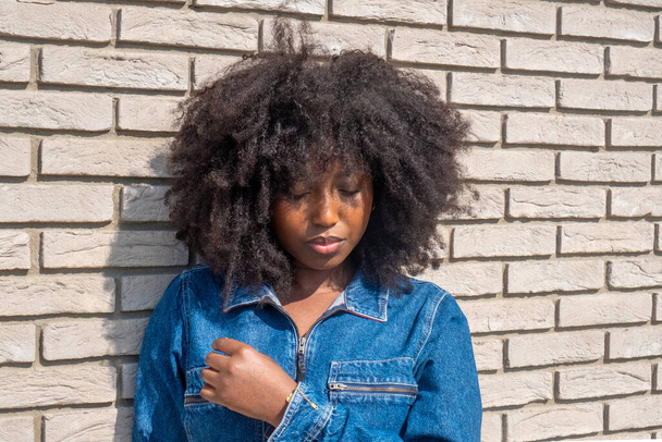 Захоплена в роздумах, молода афроамериканка з повною, природною афро стоїть на стіні суворої білої цегли. Сонячне світло підсвічує її джинсову куртку, додаючи тепло до зображення, а - Фото, зображення