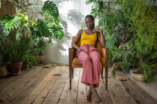Klidná mladá černoška odpočívající na židli v útulném zeleném bytě s monstera rostlin, zamyšlená Afroameričanka tráví čas, dívá se na kameru odpočívající v křesle na domácí zahradě. Bez stresu - Fotografie, Obrázek